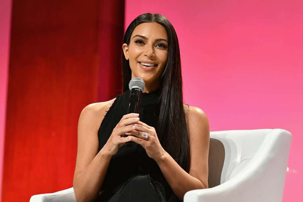 Kim Kardashian in black in front of pink background with microphone